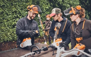 3 Menschen mit Gartentechnik im Vordergrund, die sich einen Akku der Marke STIHL ansehen. Im Hintergrund eine weitere Person mit Heckentrimmer in der Hand.