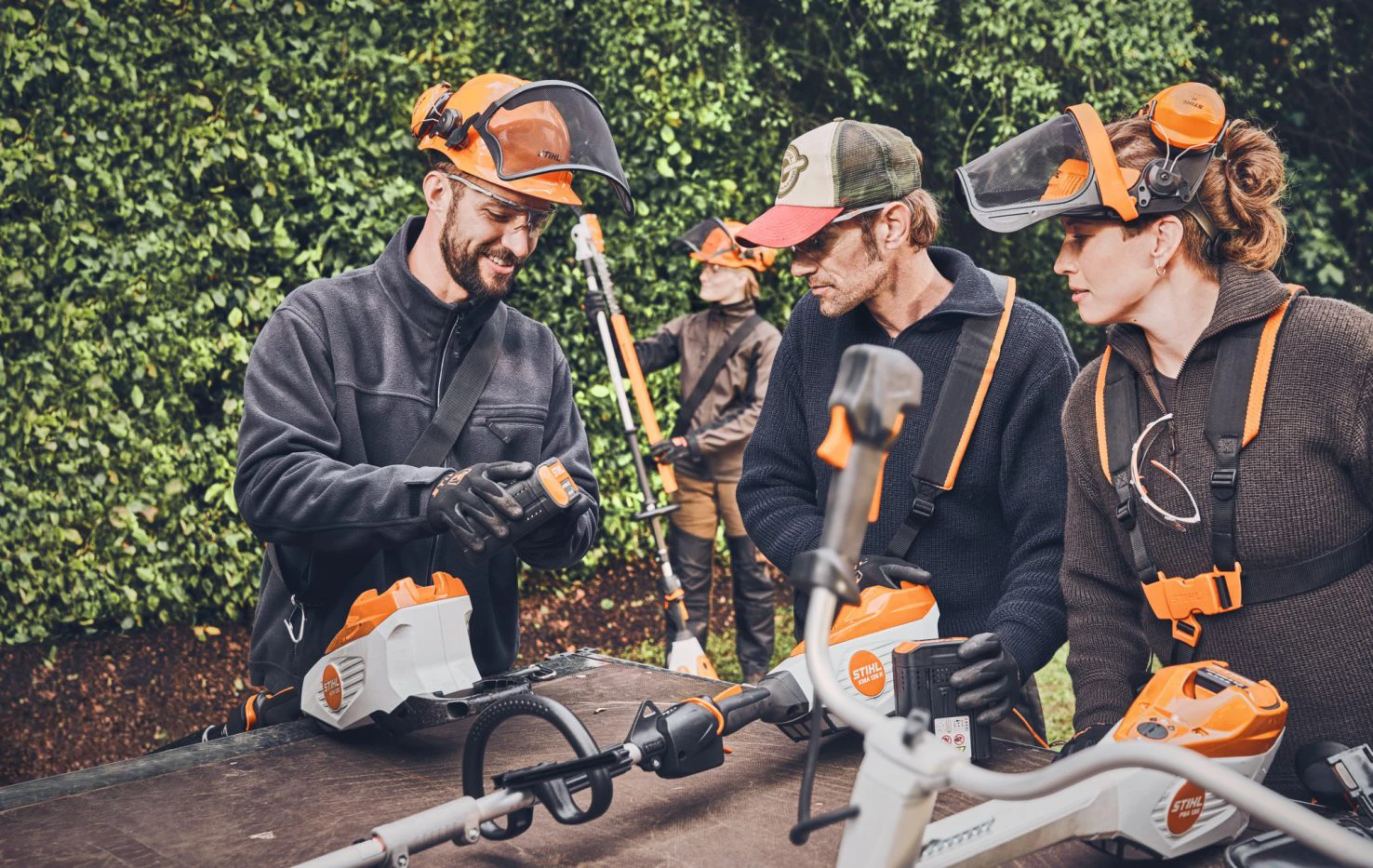 3 Menschen mit Gartentechnik im Vordergrund, die sich einen Akku der Marke STIHL ansehen. Im Hintergrund eine weitere Person mit Heckentrimmer in der Hand.